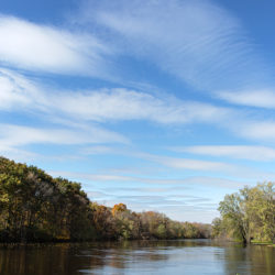 The Milwaukee RIver between wooded shorelines in autumn under a sky full of long, thin clouds