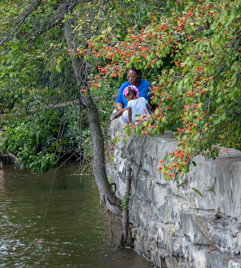 A woman and a young girl sitting on a rock wall, fishing in the Milwaukee River, with red berries overhanging them