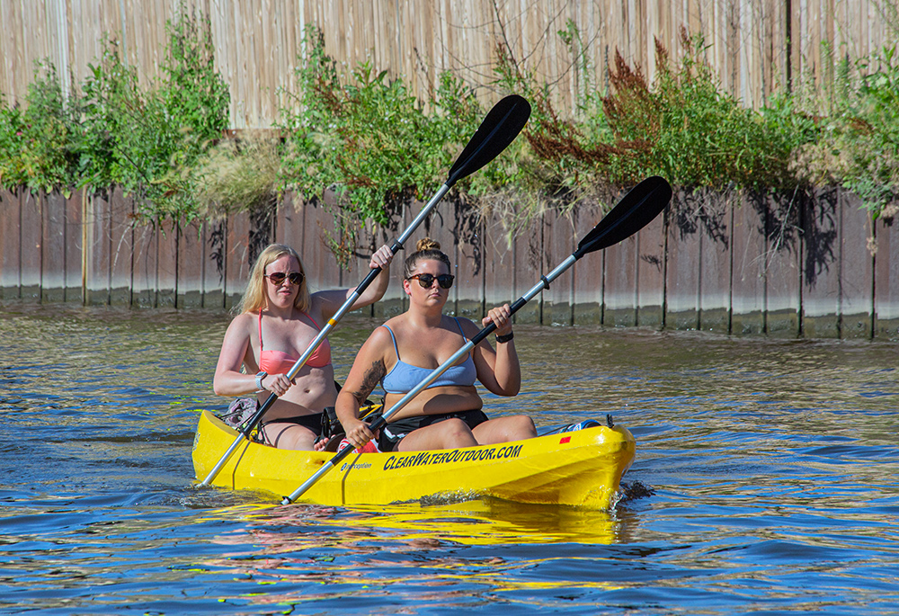 two young women in a two-person kayak paddling next to a steel floodwall on the Milwaukee River.