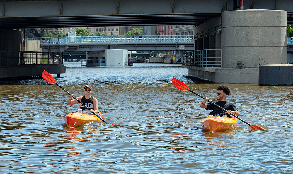 two kayaks paddling in tandem with bridges in the background on the Milwaukee River.