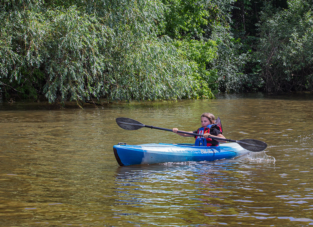 a boy in a kayak with a large paddle next to a wooded shoreline on the Milwaukee River.