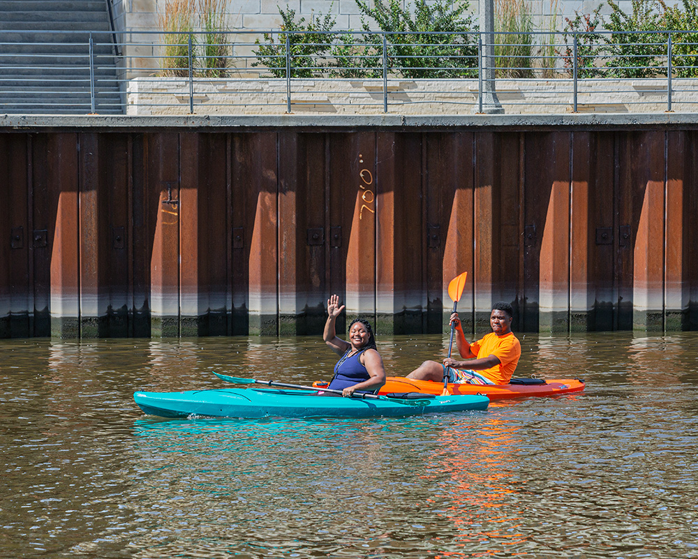 two kayakers, one waving, paddlng next to a steel flood wall on the Milwaukee River.