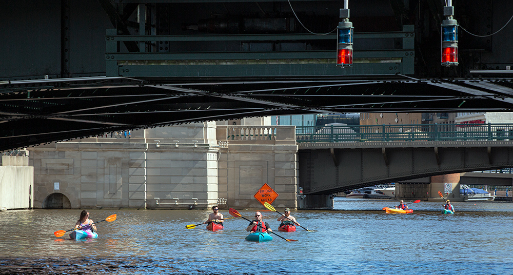 six kayaks appear tiny below the bridges of downtown Milwaukee on the Milwaukee River.