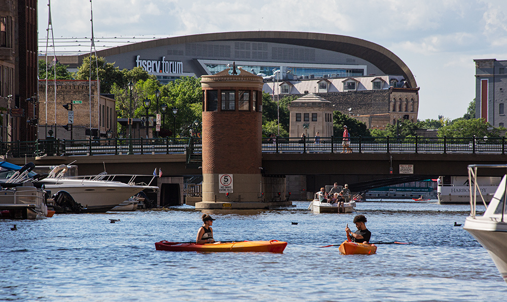 two kayaks and some motorboats on the Milwaukee River in downtown Milwaukee with the Fiserv Forum in the background