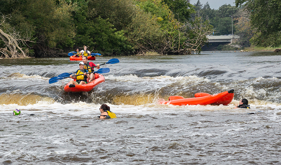 a raft going over the falls on the Milwaukee River as another raft waits and one is capsized under the falls