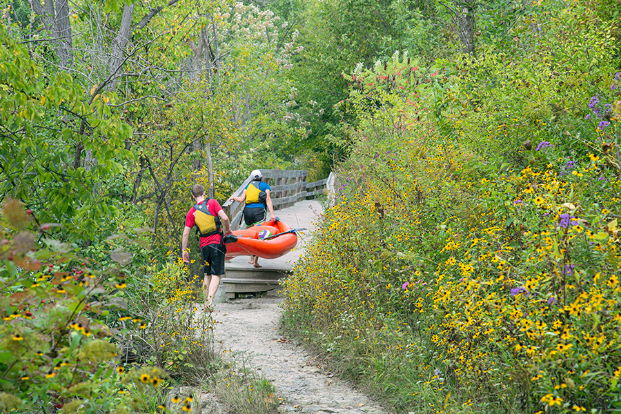 two young people portaging a red rubber raft on a boardwalk trail alongside the Milwaukee River