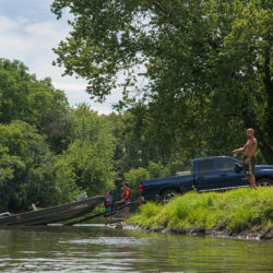 a man fishing next to a boat being pulled out of the river by a pickup truck