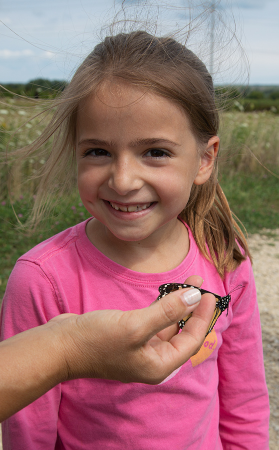 a young girl with a smile as a hand holds out a monarch butterfly