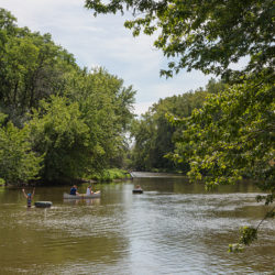 people with a canoe and rafts playing in a river with wooded shores