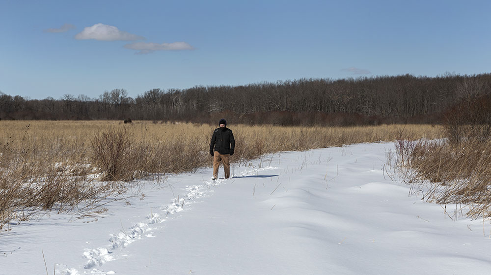 A man walking on a wide, snowy path in an open prairie at Fellenz Woods Preserve