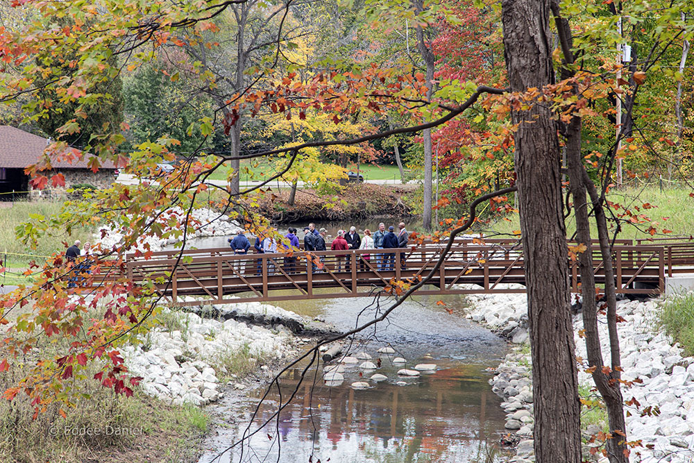 Autumn colors frame the group of dignitaries and citizens congregating on a newly installed pedestrian bridge over the Pike River.