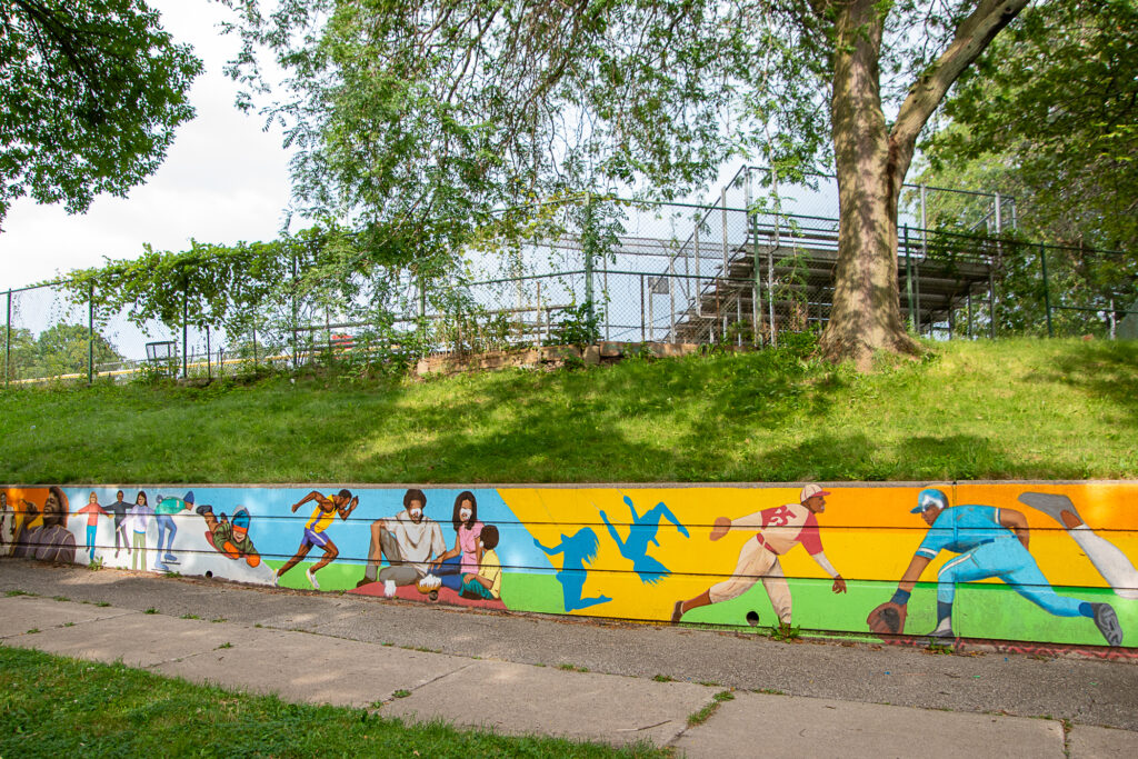 A mural depicting athletics and other recreational activities on a park retaining wall. KGM