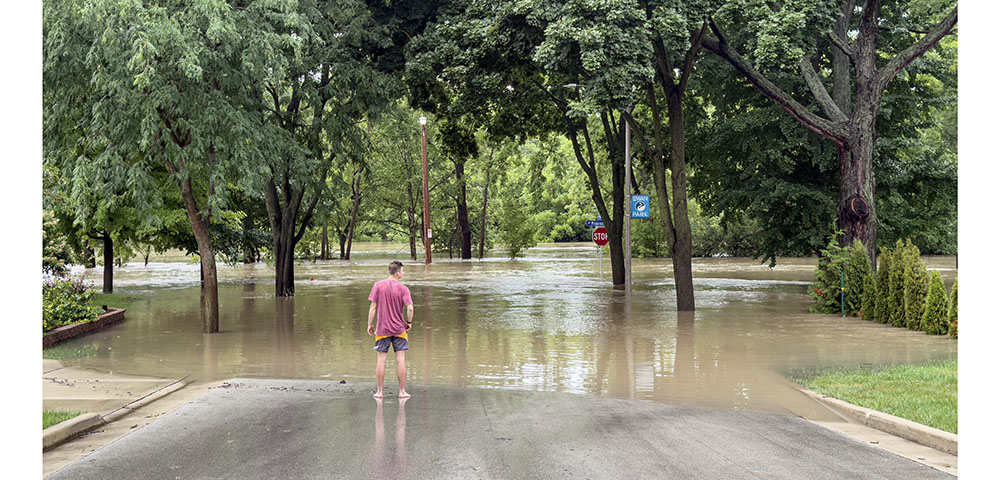 A man stands before the flooding Menomonee River at the end of a street dipping into water.