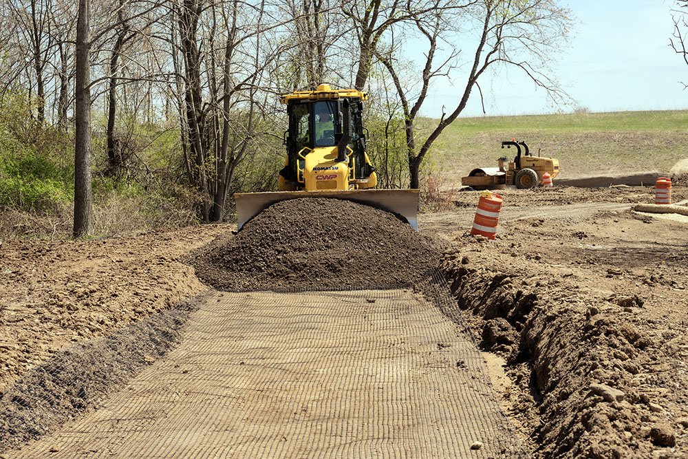 The finished trail is a layered construction. First a shallow trench is dug. Then a reinforcement mesh layer is laid on top, followed by gravel, which is being pushed into place by a bulldozer. The asphalt is added on top of all this.